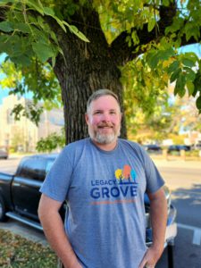 The man Jayd Raines with salt and pepper hair and beard standing in front of a tree on a beautiful sunny day. He is wearing a Legacy Grove T-shirt.