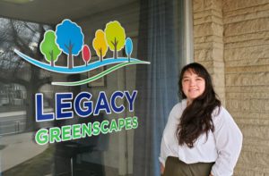 Woman standing in front of a large window with a decal logo that says Legacy Greenscapes and has different colored trees. She has long dark hair and is wearing a white long sleeve button down.